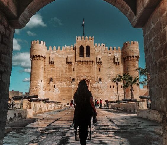 Woman walking towards a grand medieval fortress under an archway, with a clear blue sky above.