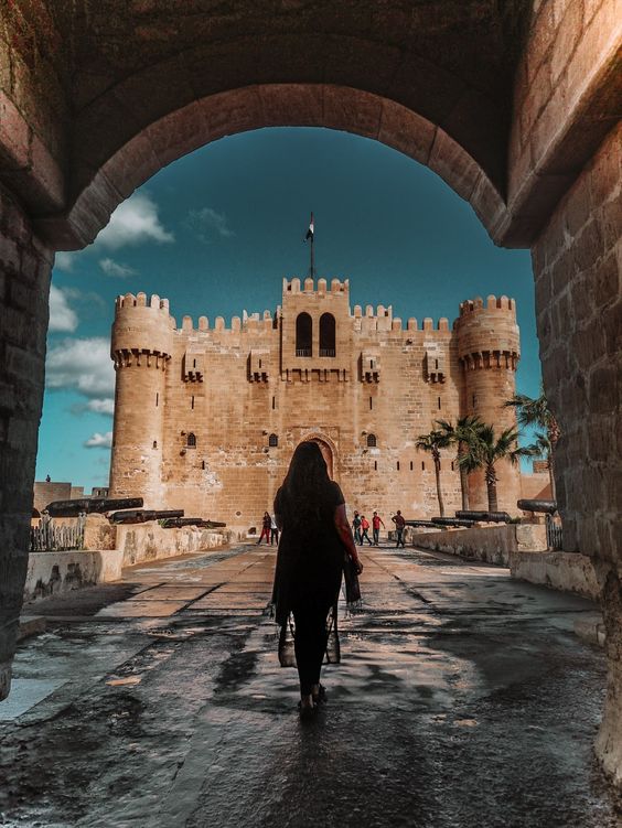 Woman walking towards a grand medieval fortress under an archway, with a clear blue sky above.