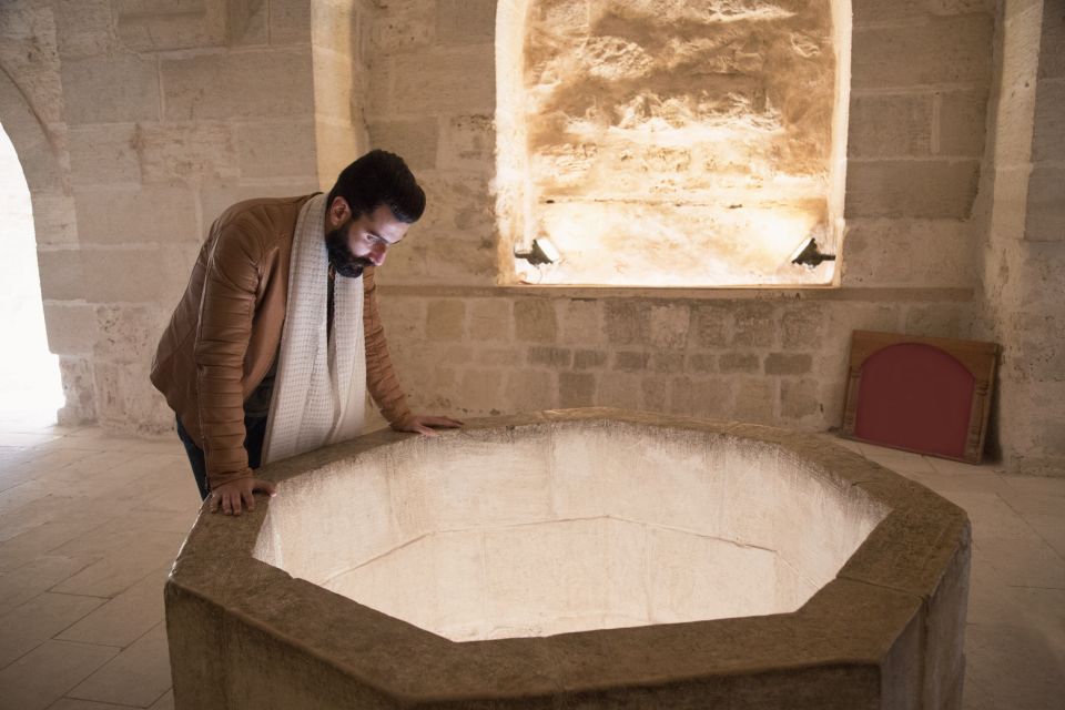 Person in a jacket and scarf leaning over a large stone basin in a room with arched windows.