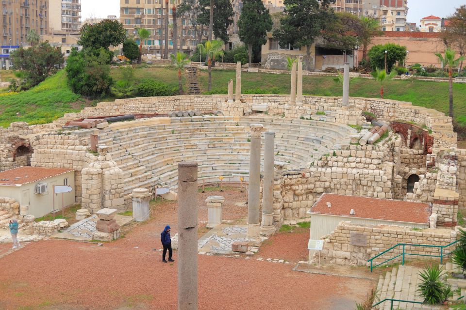 Ancient amphitheater with ruins and columns, surrounded by modern buildings, with people wandering.