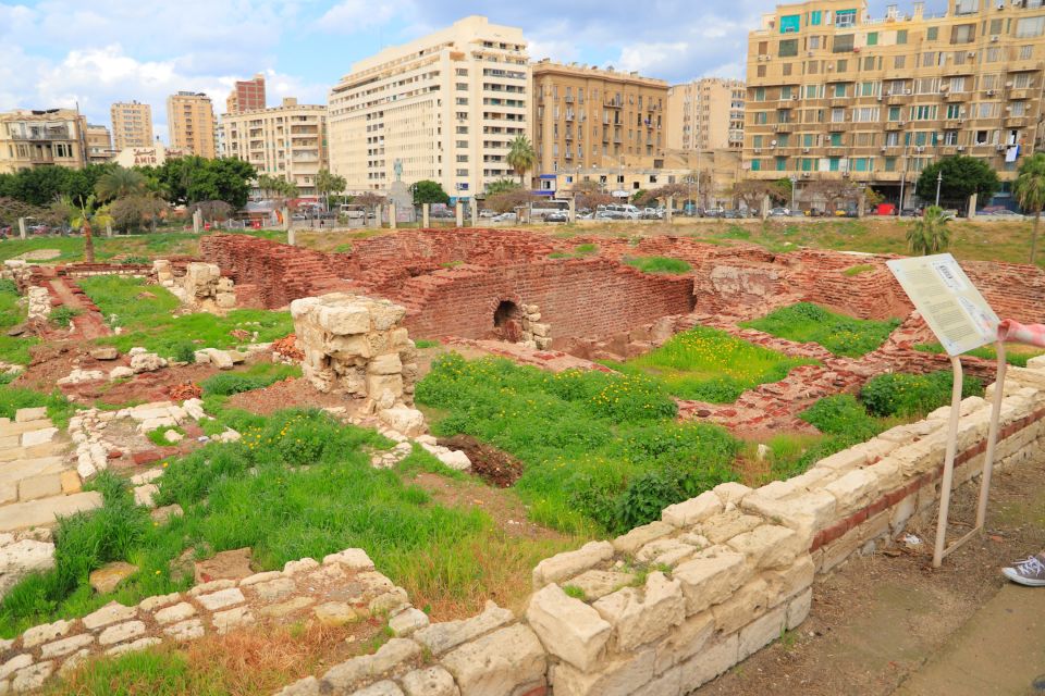 Ancient ruins in a grassy field with modern buildings in the background.