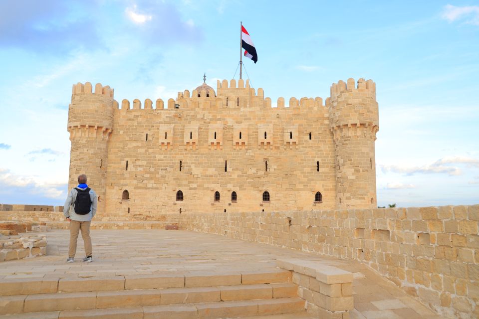 A person standing in front of an ancient stone castle with a flag on top under blue skies.