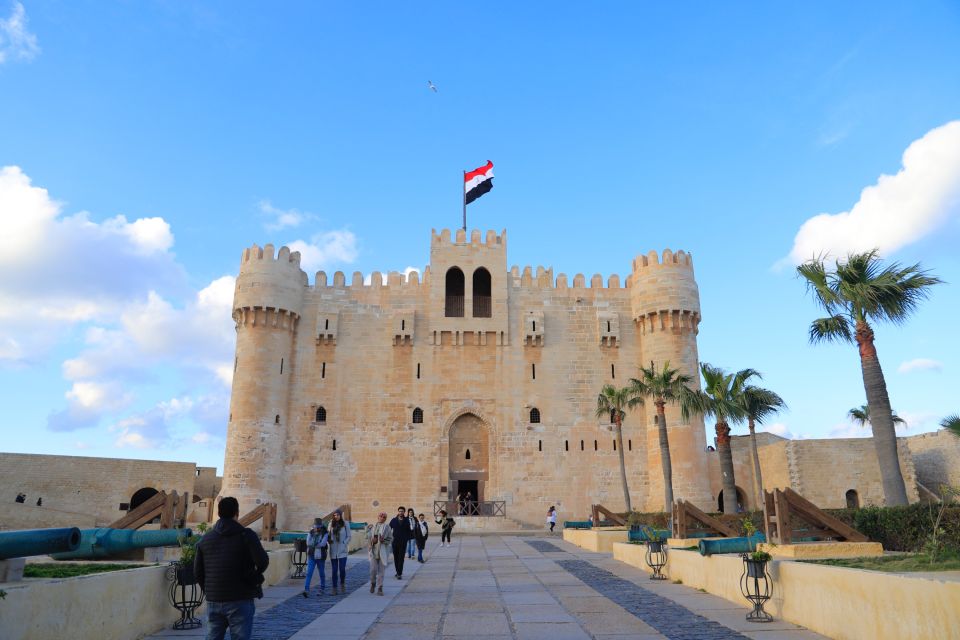 A majestic stone fortress with a flag on top under a blue sky, flanked by palm trees and cannons.