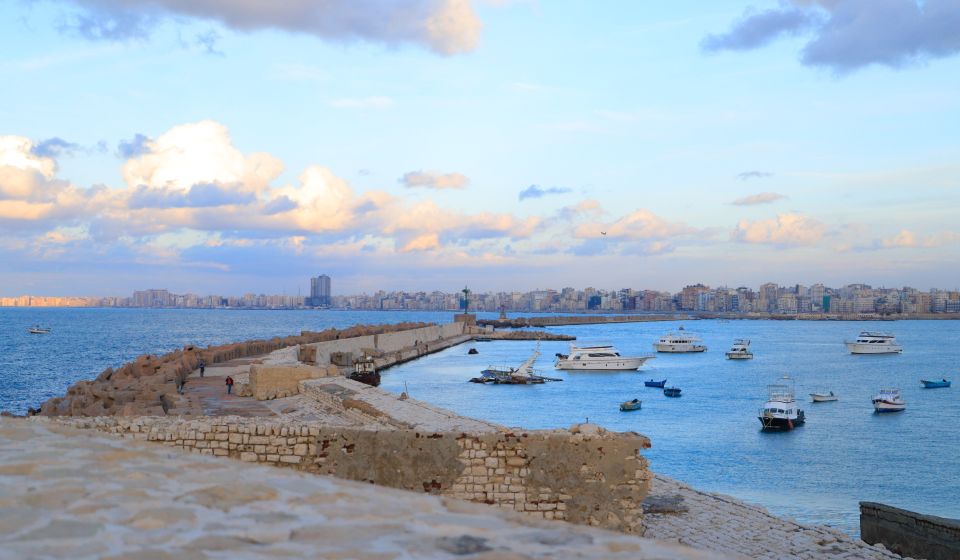 Seaside cityscape at dusk with boats and ancient stone pier under a cloudy sky.