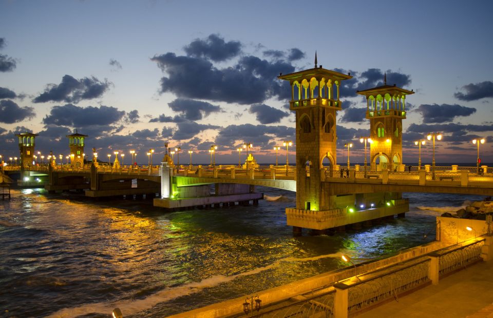 Night view of the illuminated Stanley Bridge in Alexandria, Egypt, with glowing streetlights and a partially cloudy sky.