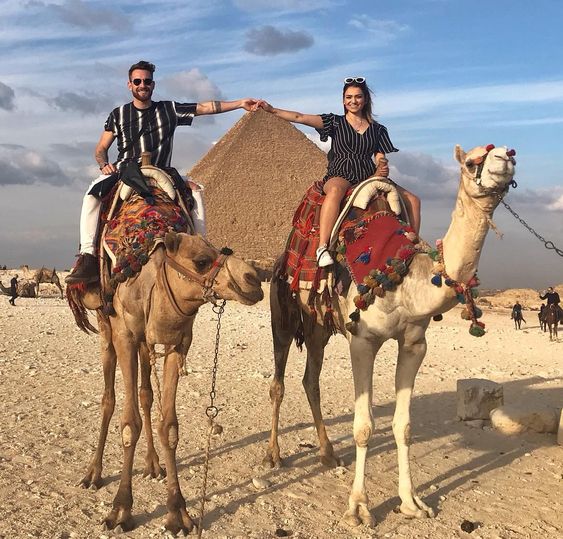 Two people riding camels in front of the Great Pyramid of Giza, under a blue sky.