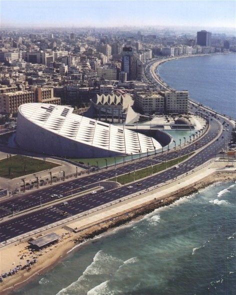 Aerial view of a curved building along a coastal road with a dense cityscape in the background.