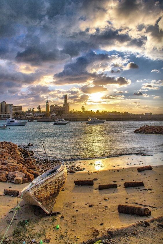 Sunset over a harbor with boats, a beached old boat in the foreground, and a city skyline with clouds.