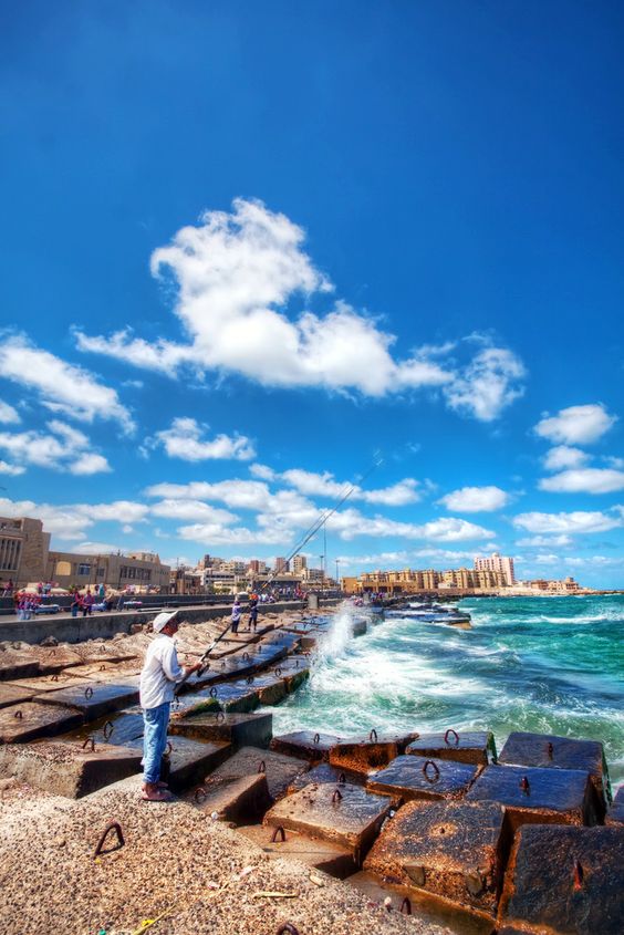 People fishing on a jetty with waves crashing and a bright blue sky with clouds.