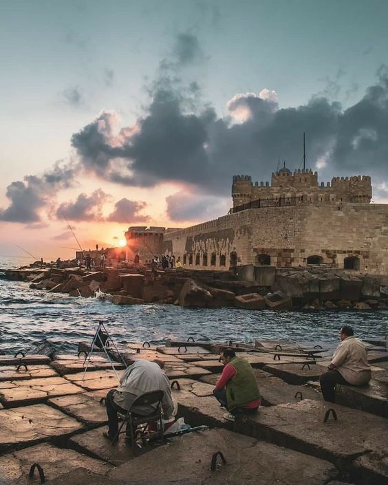 Sunset behind a fortress with people fishing on the shoreline.